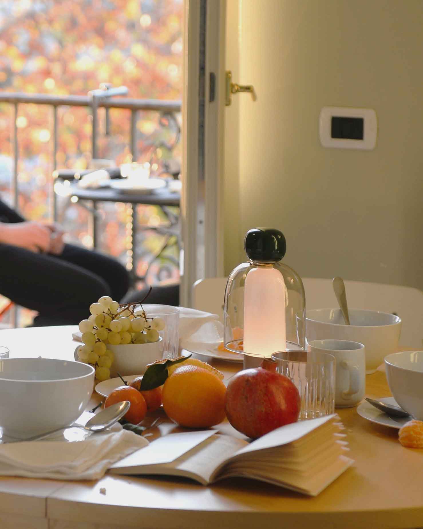 [Appartamenti] Dettaglio colazione, libro, arancia e melagrana in primo piano rispetto al balconcino, fuori fuoco, dove prendere l'ancora caldo sole autunnale di Roma. [Appartamenti] Breakfast detail, a book, orange, and pomegranate in the foreground, with the small balcony out of focus, where you can catch the still-warm autumn sun of Rome.